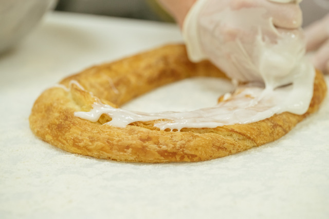 Two female bakers with aprons, hairnets, and gloves creating kringle with dough smile as they work