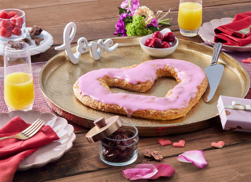 Heart-Shaped Kringle with pink icing surrounded by Valentine's Day decorations. 