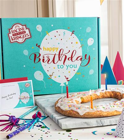 Turquoise box with "happy birthday to you" type behind a Kringle on a marble board surrounded by birthday horns, hats, and confetti. 