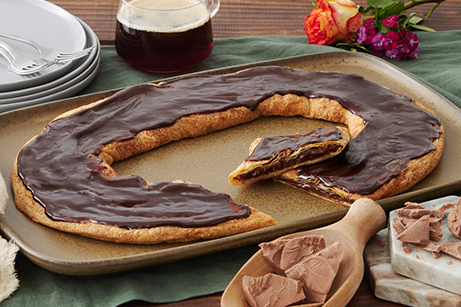Chocolate Silk Kringle of white plate with slice on smaller plate surrounded by bowl of chocolate and butter stick. 