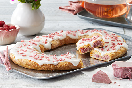 Ruby Chocolate Raspberry Kringle on a silver tray surrounded by a bowl of raspberries, chunks of ruby chocolate, pitcher of pink colored tea and vase of flowers. 