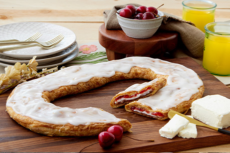 Cherry Cheesecake Kringle on white plate with slice cut on on small plate surrounded by bowls of cherries, flour and eggs. 