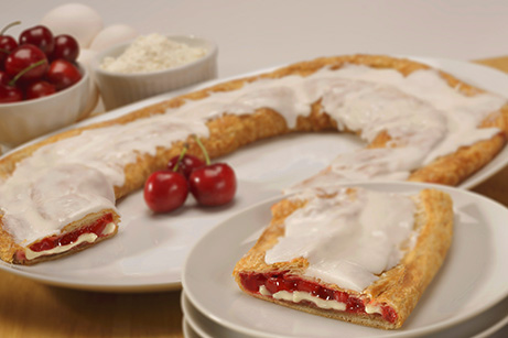 Cherry Cheesecake Kringle on white plate with slice cut on on small plate surrounded by bowls of cherries, flour and eggs. 