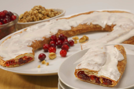 Cranberry Nut Kringle on a white plate with a slice on a smaller plate along with bowls of cranberry and walnuts.