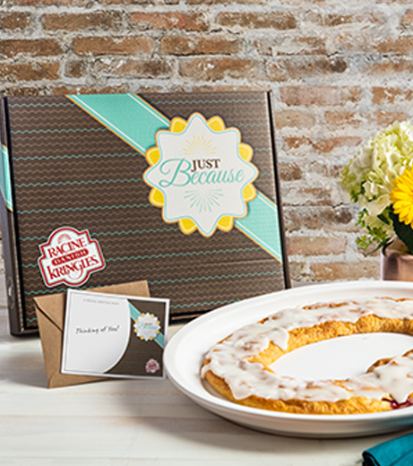 Brown box with turquoise stripe with "Just Because" font behind a kringle on a white plate next to a vase of yellow flowers and a brick wall background.