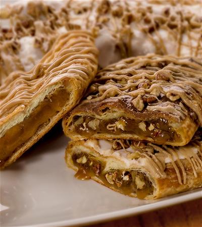 Praline Pecan Kringle with slices of Turtle Kringle and Sea Salt & Caramel Kringle with bowls of chocolate blocks, cubed caramels and pecans. 