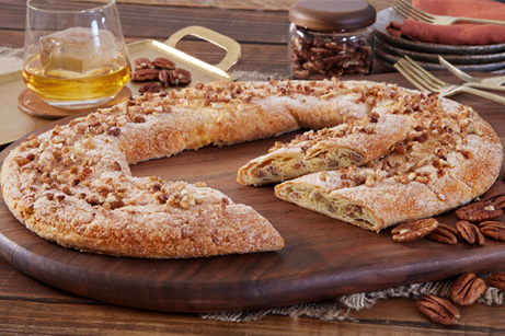 Bourbon Pecan Kringle on a white plate with a few pecans and a glass on bourbon next to Kringle. 