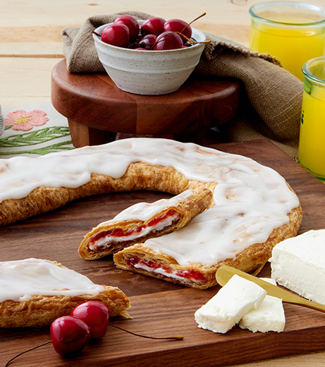 Cherry Cheesecake Kringle on white plate with slice cut on on small plate surrounded by bowls of cherries, flour and eggs. 